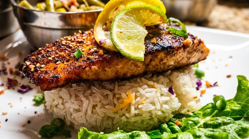 Grilled salmon with sesame seeds, lime, and lemon slices, served over rice with lettuce.