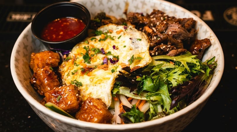 A colorful bowl of food with beef, fried egg, glazed tofu, mixed greens, and a side of red sauce.