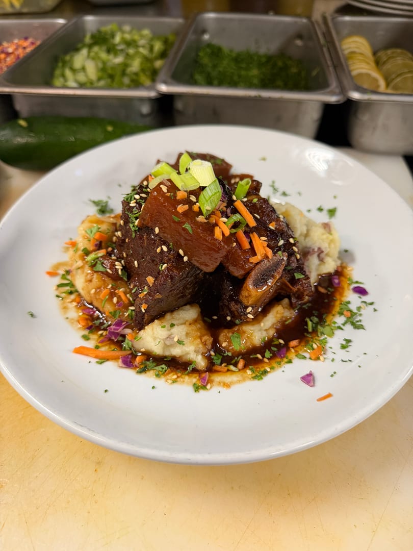 Braised short ribs on mashed potatoes, garnished with scallions, carrots, sesame seeds, and a rich sauce.