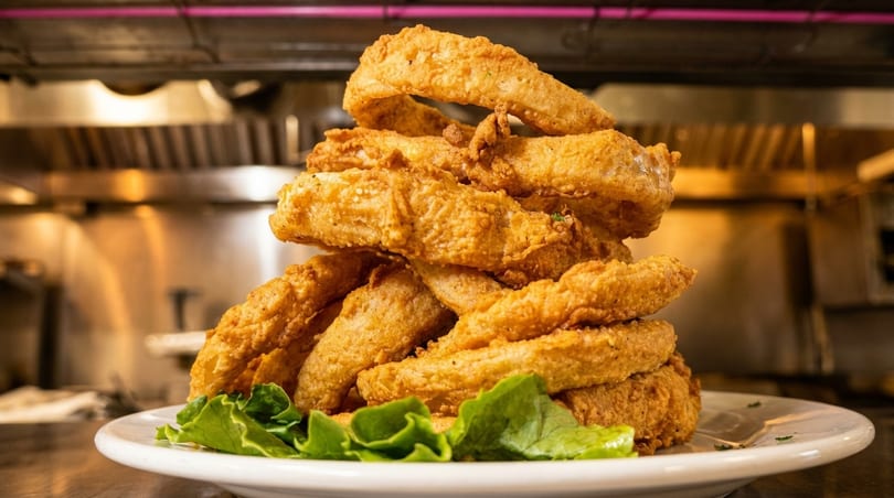 A towering stack of golden-brown fried onion rings on a white plate with lettuce, in a restaurant kitchen.